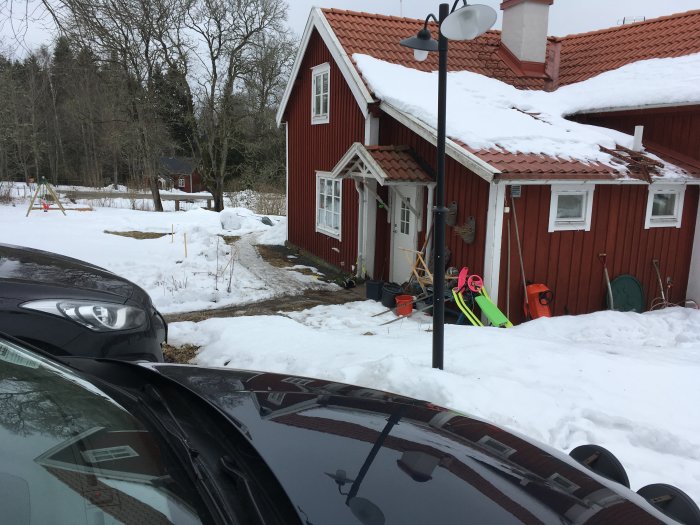 Röd stuga med snötäckt tak och gavlens del där en utbyggnad planeras, omgiven av snö och trädgårdsredskap.