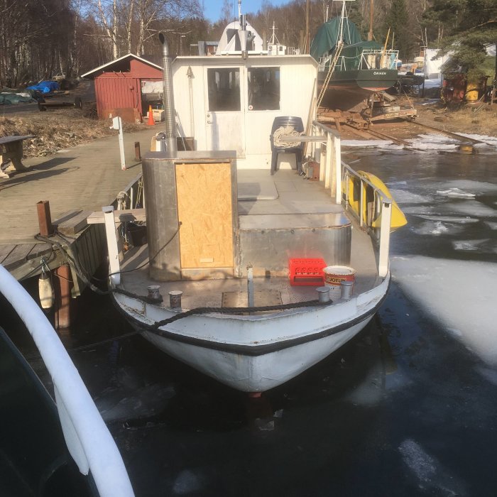 Båt med provisorisk plywooddörr och pågående reparation, förtöjd vid isig brygga.