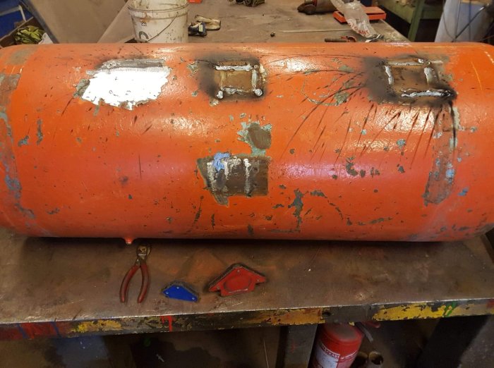 En rostig orange gasflaska med flera nyligen svetsade lappar på ytan. Verktyg syns på arbetsbänken framför.
