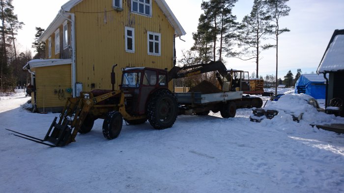 Gul traktor med frontlastare och släpvagn lastad med jord framför gult hus i snöigt landskap.