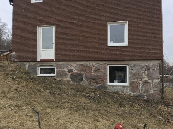 Plats för planerad altan med lutande mark och vegetation framför tegelhus.