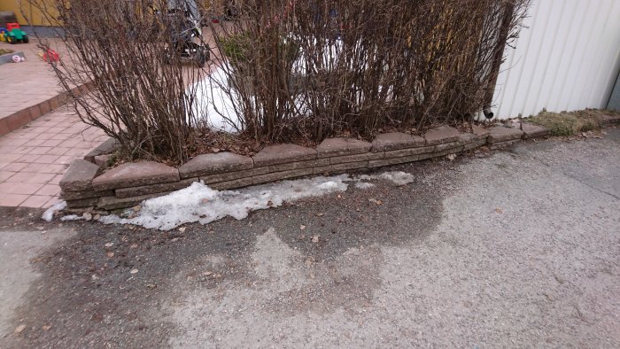 Otillfredsställande installation av kantstenar vid en rabatt med buskar och smältande snö.