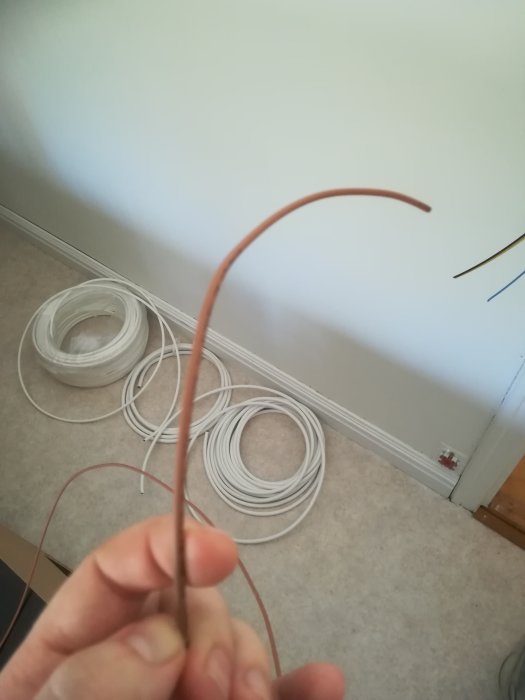 En hand håller en brun tråd med en rulle vit kabel i bakgrunden på ett ljust golv.