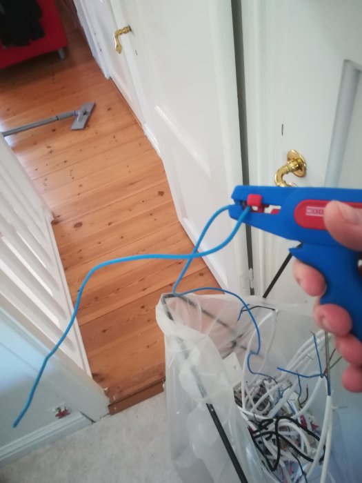 En hand håller en blå skaltång som böjer en blå elkabel i ett hemmiljö med trähögolv.