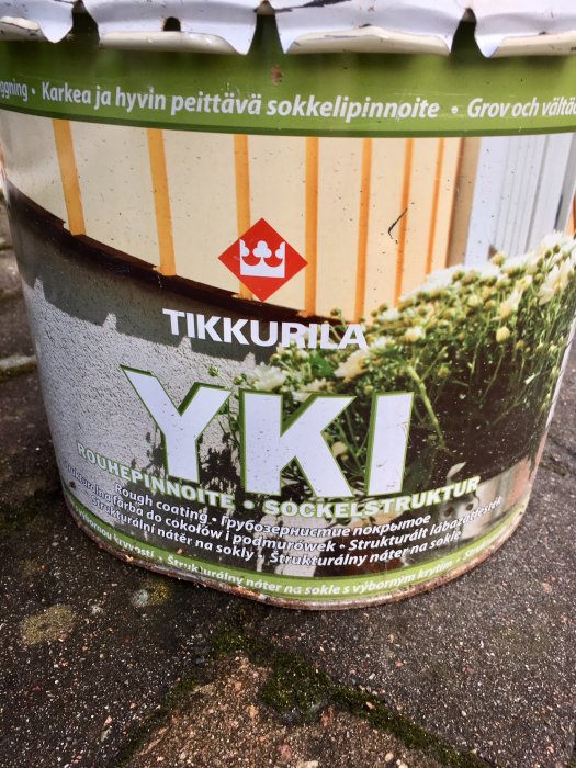 En använd burk av Tikkurilla Yki Sokkelistructuur, en grov putsprodukt, står på en stenplatta med mossa.