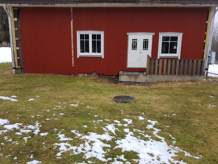 Röd vägg på en stuga med två fönster och en dörr, träveranda, brunnen nära huset på en gräsmatta med snöfläckar.