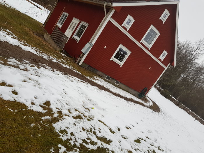 Rött trähus med snötäckta gräsytor i förgrunden och nakna träd i bakgrunden.