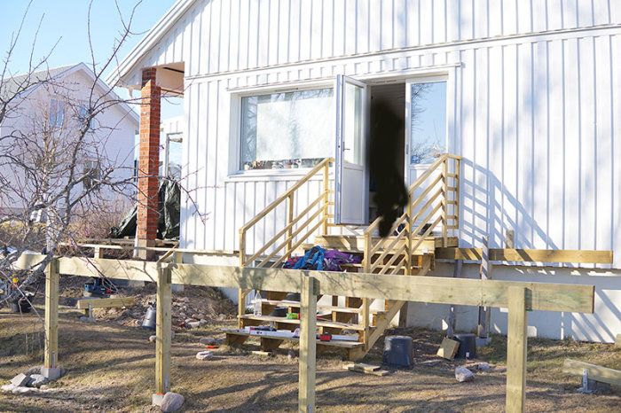 Träkonstruktion av bärlina och stolpar vid hus, verktyg och byggmaterial synliga på marken.