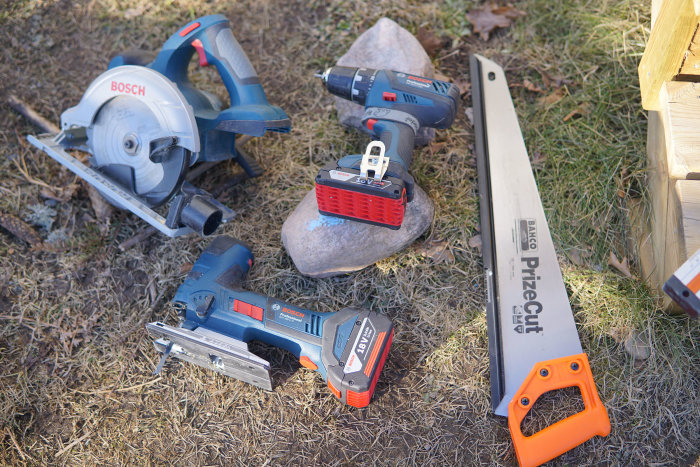 En samling verktyg på marken inklusive en Bosch cirkelsåg, borrskruvdragare, batteripack och handsåg, antyder nötning vid byggarbete.