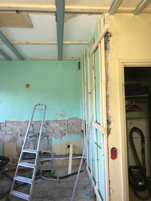 Ett hörn av ett pågående byggprojekt med osymmetrisk slaggbetongvägg och intilliggande reglar för kommande OSB och gipsbeklädnad.