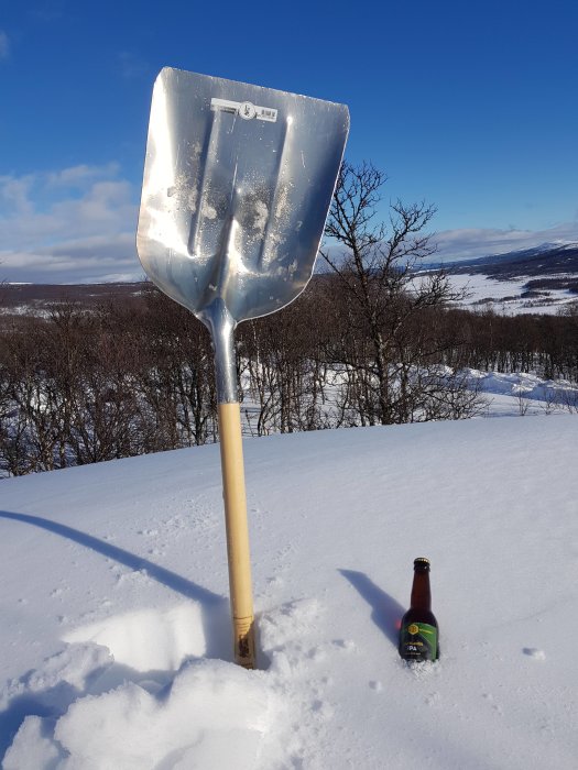 En snöskyffel står vertikalt i snön intill en flaska öl, mot en bakgrund av snötäckt landskap och blå himmel.