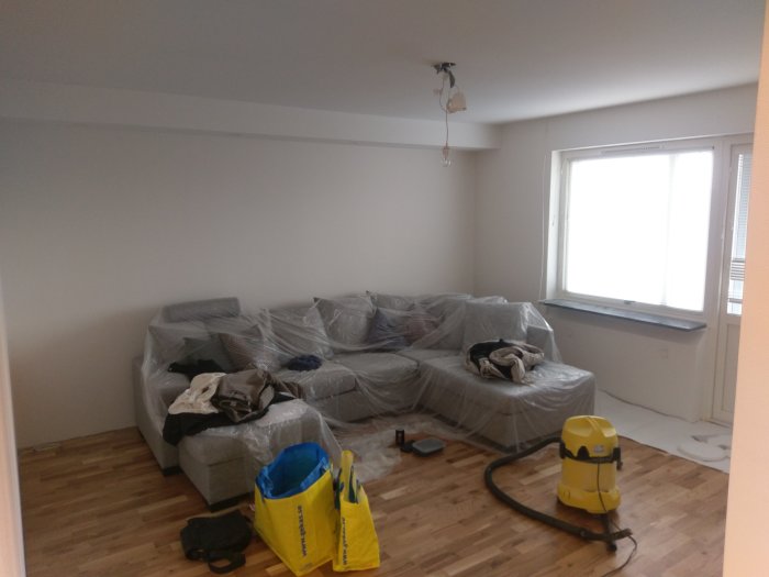 Vardagsrum med soffa skyddad av plast, avtäckt golv och målarutrustning under renovering.