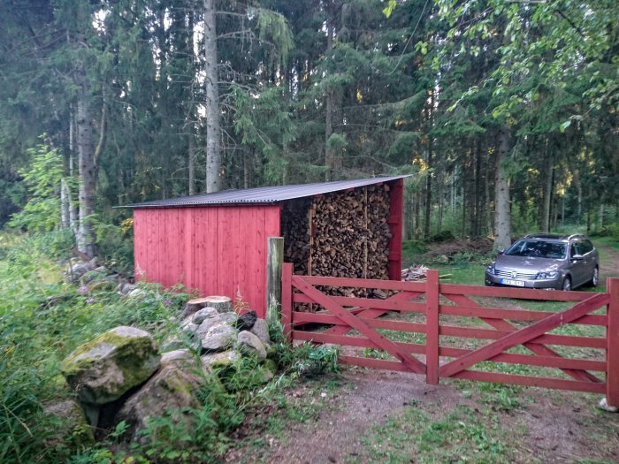 Rödmålad träförrådsbyggnad med öppen vedförvaring och plåttak, belägen i skogsmiljö intill en grusväg och ett rött staket.