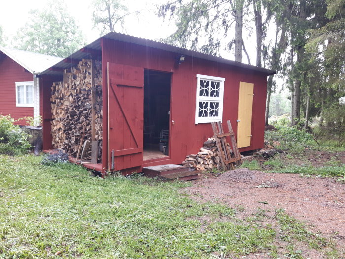 Träförråd med röda väggar och gult dörr, halvtäckt vedstapel vid sidan, nära grönt hus.