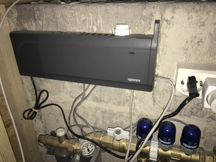 En Uponor-fördelarskåp med röranslutningar och blå rattar, mätenhet och elsladdar mot betongvägg.