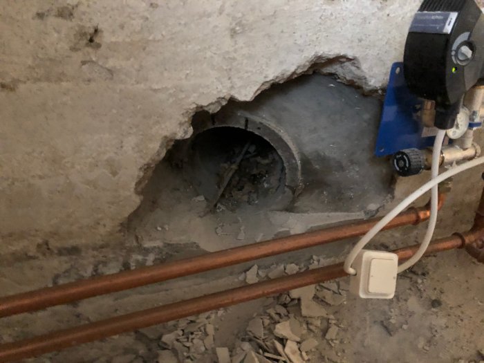 Hål i källarvägg där en gammal kulvert har varit, omgivet av rör och en pump, behöver igenmurats och putsas.