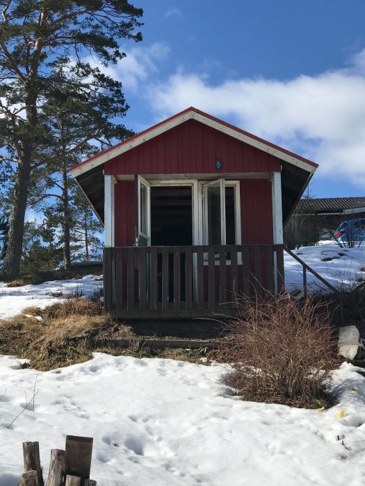 Röd gäststuga på 8kvm med trädäck framför i vintersol, snö runtomkring.