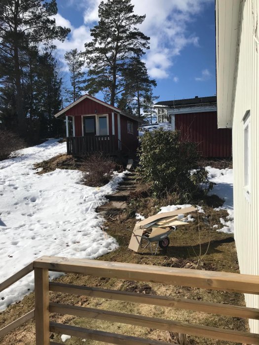 Liten röd stuga på snötäckt tomt med träd i bakgrunden, synlig från en trätrappa.