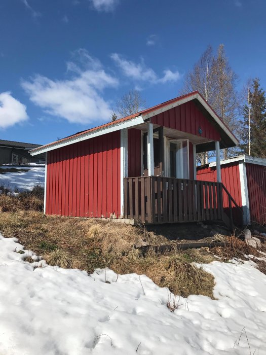 En liten röd gäststuga med vit vidkanten står på en gjuten platta omgiven av smältande snö.