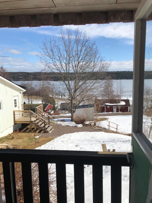Utsikt från gäststugans veranda med snötäckt mark och en sjö i bakgrunden, omgiven av träd och byggnader.