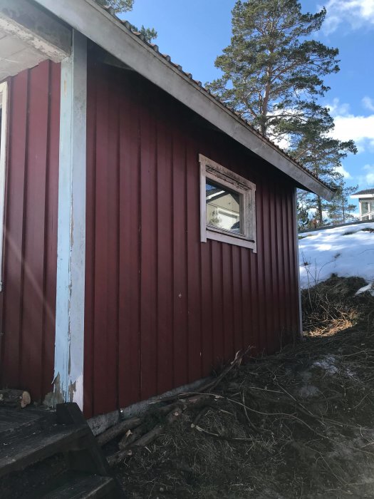 Rödmålad gäststuga med flagnande färg på panelen, ett litet fönster, och synlig grund på en gjuten platta.