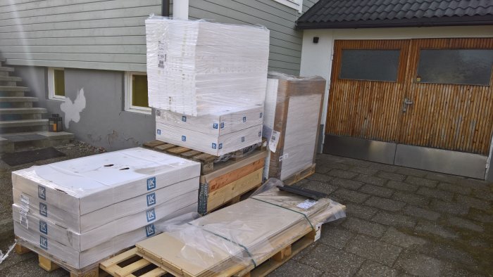 Pallar med förpackade golvvärmekomponenter framför hus med träfasad och garageport.