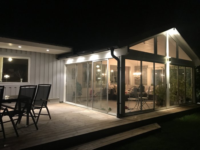 Upplyst uterum med glaspartier på träterrass kopplat till vit husfasad på kvällen.