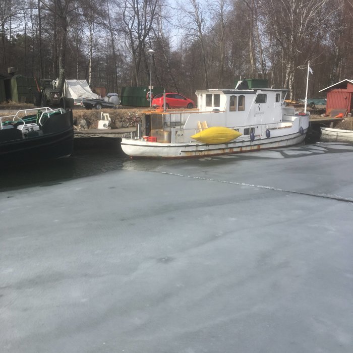 Båtar i en frusen hamn med isbelagd yta och skog i bakgrunden.