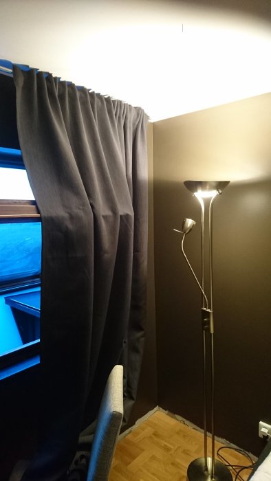 Mörkt sovrum med bruna väggar och grå gardiner i hörnet, golvlampa och delvis öppet fönster.