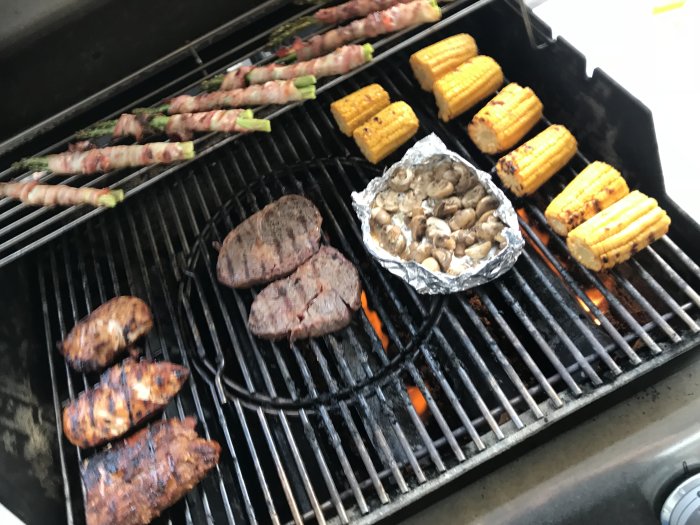 Grillat kött, majskolvar och baconlindade sparris på en tänd grill.