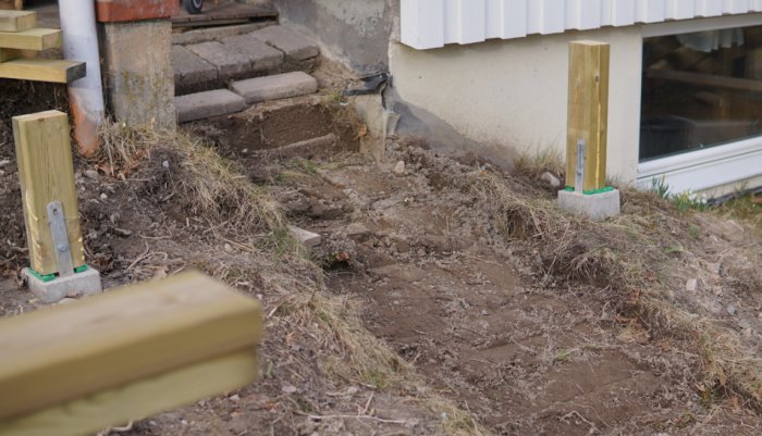 Pågående byggprojekt med nyinstallerade stolpar och skyddshinkar vid husgrund, tidigare stentrappa syns.