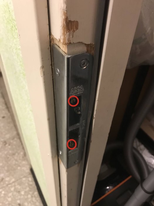 Svart vinkelslutbleck inbyggt i dörrkarm med två markerade skruvhål, nära en skadad karmkant.