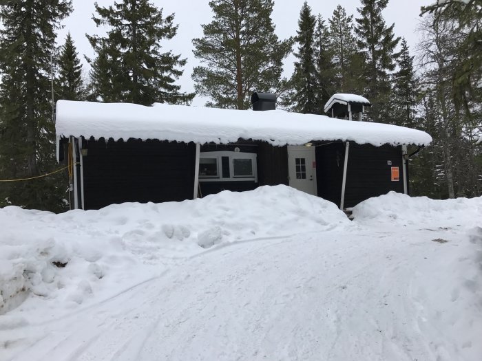 Svart timmerstuga med tjockt snötäcke på taket omgiven av snödrivor och skog.