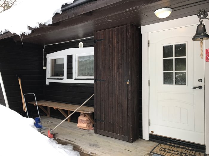 Entré till mörkt trähus med vit dörr, fönster och snöskottning verktyg på verandan under vintern.