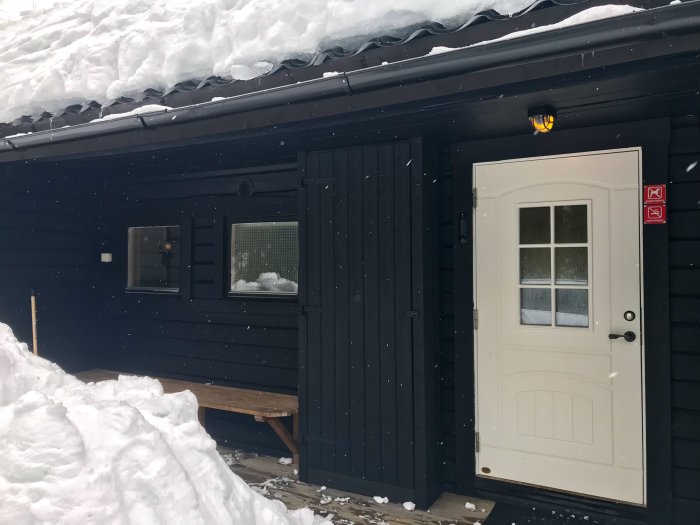 Svartmålad stuga med vita detaljer under en snötäckt takkant, inklusive en dörr, fönster och skidförvaring bredvid dörren.