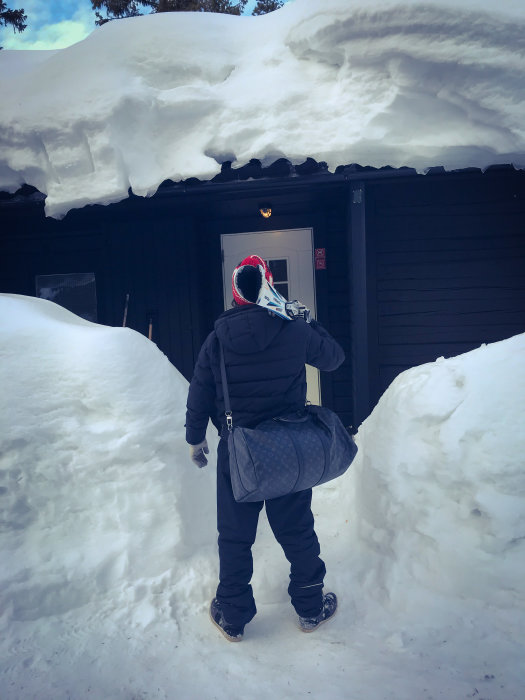 Person står framför en dörr till en stuga täckt av tjockt snötäcke i Lindvallen.