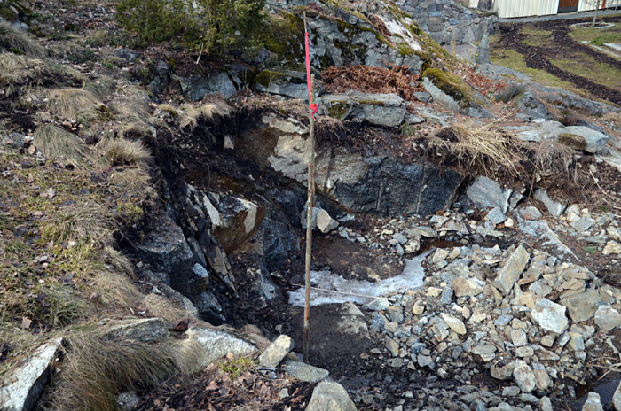 Sprängd berggrund i en sluttning med mätstolpar och röd linje som markerar framtida växthusgavel och takfotshöjd.