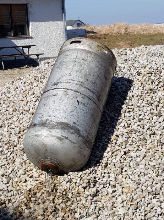 En liggande gasoltank som töms på vatten på en grusplan framför en byggnad.