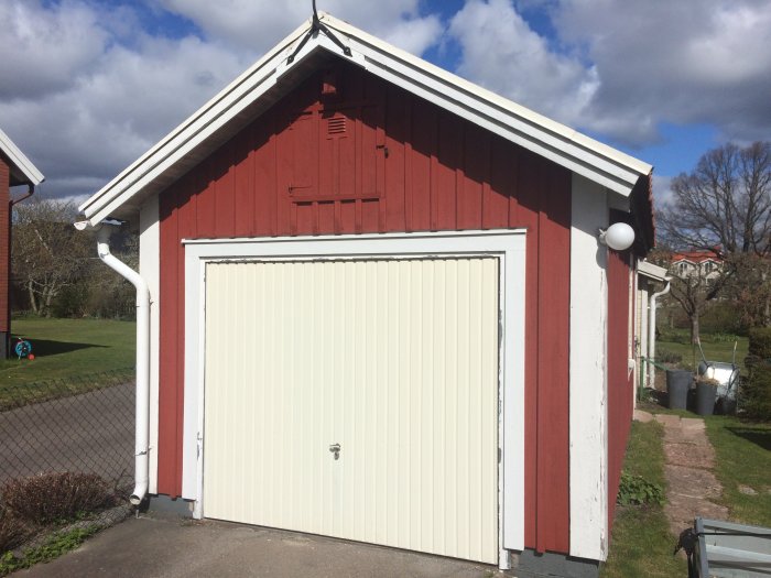 Röd verkstad med vit garageport, rännor och ny vindflöjel i solljus med grön gräsmatta i bakgrunden.