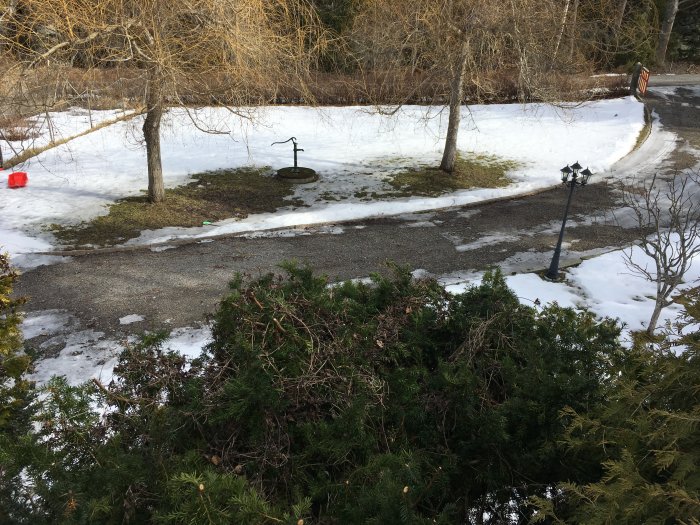 Platt tomt med stående vatten på grus och gräs, snöpartier, och träd vid brunnen.