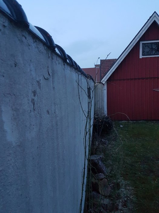 Sliten grå betongmur med taggtråd intill en röd stuga och stenmur vid skymning.