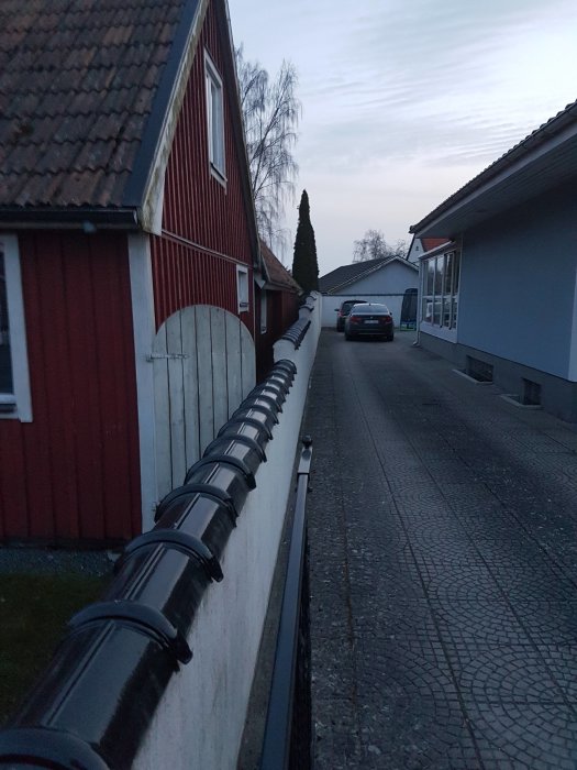 En förgård med tegeltakhus, staket och svart stuprör längs en trottoar vid skymning.