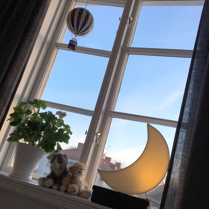 Ett mysigt fönster med en söt månformad lampa, två gosedjur och en grön krukväxt med utsikt mot himlen.