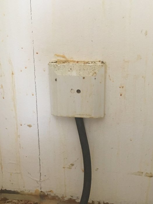 Äldre vägguttag med sliten yta och en svart elkabel ansluten, monterat på en vit vägg med fläckar.