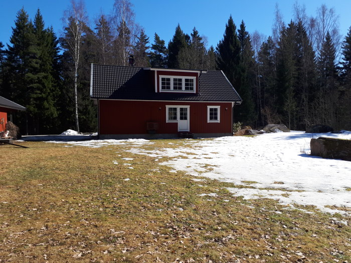 Ett rött hus med vita knutar och svart tak på en snötäckt gräsmatta med skog i bakgrunden.