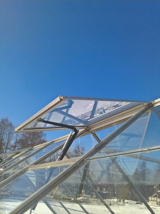 Öppen vädringslucka på ett växthus en solig dag med snön som smälter runtomkring.