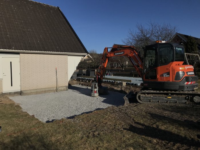 Grävmaskin intill hus med nygrävt område för dränering, singel och dräneringslock synligt.