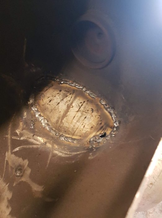 Svetsade skarvar runt bränt hål i metallyta, visar grovarbeten på en eldlåda.