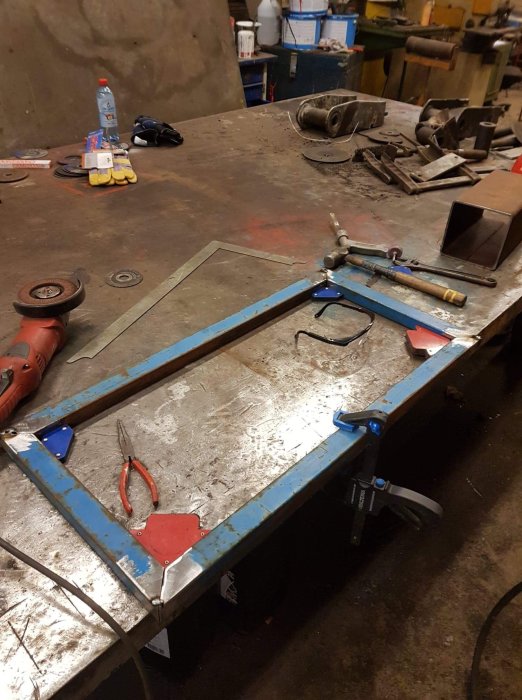 Svetsad metallram och verktyg på ett arbetsbord i en verkstad med diverse material i bakgrunden.