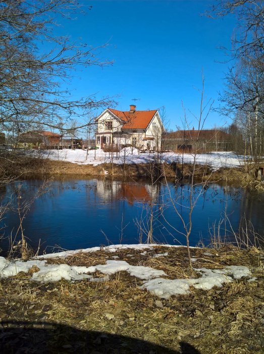En villa vid en flod med smältande snö och bar mark under en klarblå himmel.